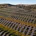 Aerial view of solar panels arranged in rows on a hillside with wind turbines in the background. - Olive Oil Times