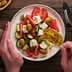 Hands pouring olive oil over a salad with tomatoes, cucumbers, olives, and feta cheese on a plate. - Olive Oil Times