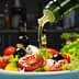 Olive oil being poured over a fresh salad with tomatoes, lettuce, and olives in a bowl. - Olive Oil Times