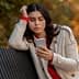 A woman with long dark hair wearing a light coat and red sweater, looking at her smartphone while sitting on a bench. - Olive Oil Times