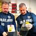 Two men in uniforms holding a metal container and a plastic bottle filled with oil. - Olive Oil Times