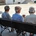Three elderly men sitting on a bench, viewed from behind, in a public square. - Olive Oil Times