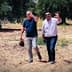 Two men walking through an olive grove, one waving and the other shielding his eyes from the sun. - Olive Oil Times