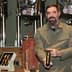 Man with a beard holding a bottle of olive oil in a production setting with equipment in the background. - Olive Oil Times