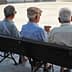 Three elderly men sitting on a park bench, viewed from behind, wearing hats and light-colored shirts. - Olive Oil Times