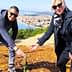 Two individuals bending down to plant a small tree in a rural area with a view of distant hills and water. - Olive Oil Times