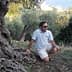 Ein Mann mit Sonnenbrille und hellem Hemd kniet neben einem Olivenbaum in einer natürlichen Umgebung. - Olive Oil Times