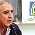Un uomo parla al microfono durante una conferenza con un logo visibile sullo sfondo. - Olive Oil Times