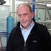 A man wearing a black jacket and a light blue shirt, standing in an industrial setting. - Olive Oil Times