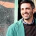 Un hombre vestido con una chaqueta verde y un suéter oscuro, sonriendo mientras está parado al aire libre cerca de una ventana. Olive Oil Times
