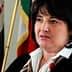 Žena tamne kose i bijele košulje govori ispred zastava. - Olive Oil Times
