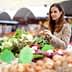 Une femme examinant des légumes frais sur un étal de marché rempli de produits divers. Olive Oil Times