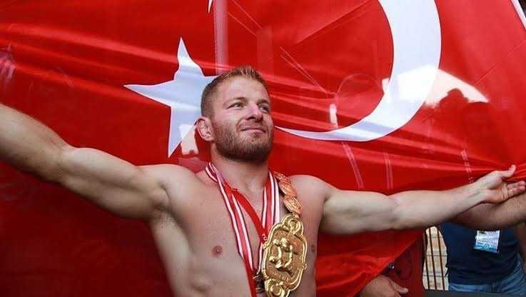 A wrestler celebrating with arms outstretched in front of a large Turkish flag, wearing a championship belt and medals. - Olive Oil Times