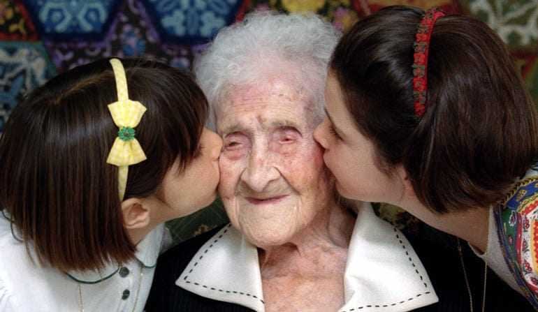An elderly woman is being kissed on the cheeks by two young girls, one on each side, both smiling. - Olive Oil Times