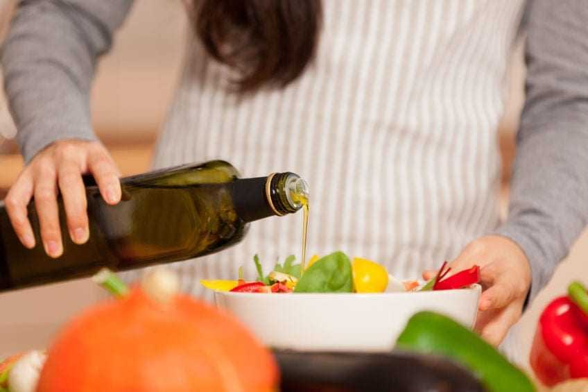 Persona que vierte aceite de oliva de una botella en una ensaladera llena de verduras. - Olive Oil Times