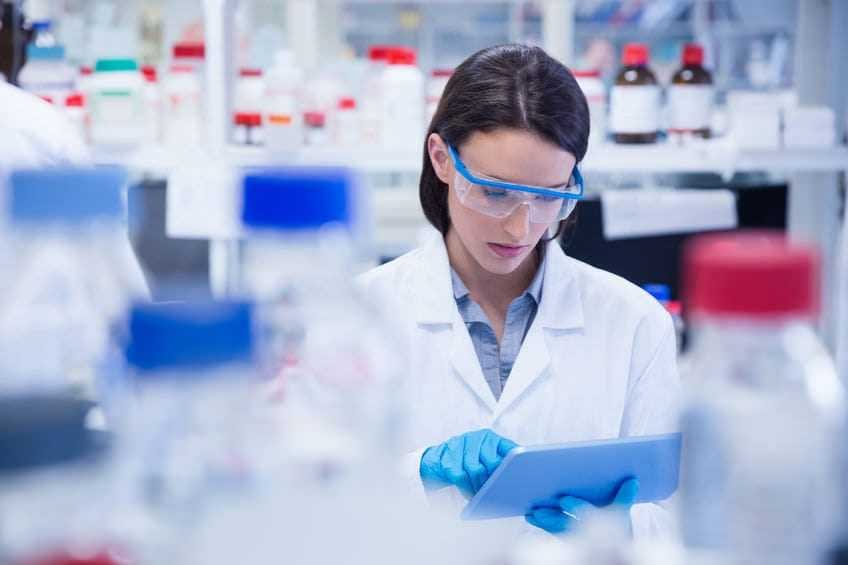 Scienziata che indossa un camice da laboratorio e occhiali protettivi mentre utilizza un tablet in un ambiente di laboratorio. - Olive Oil Times