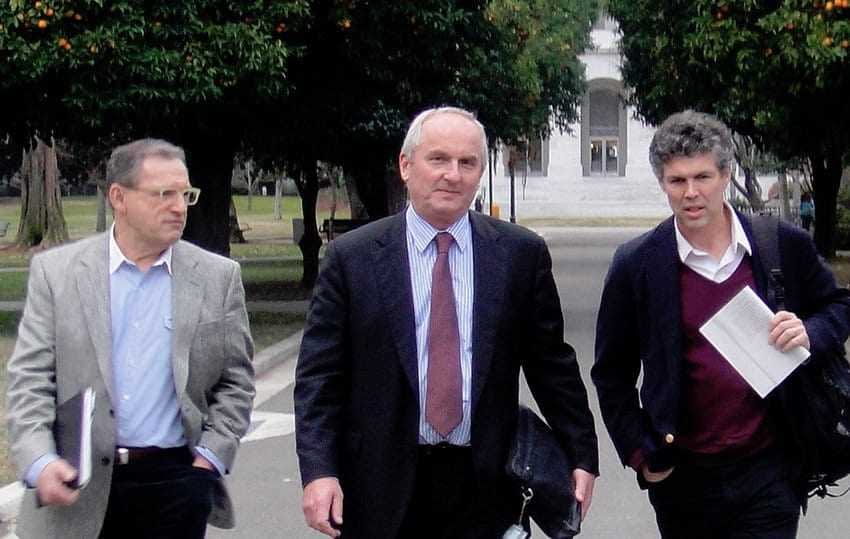 Three men walking together in a park, carrying briefcases and folders. - Olive Oil Times