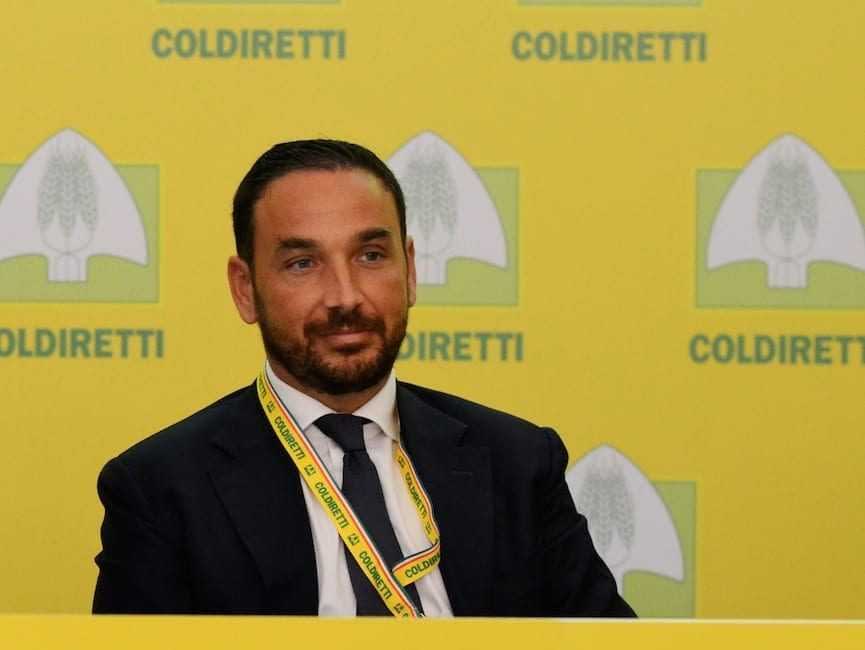 Мужчина в костюме и галстуке сидит за столом на фоне баннера Coldiretti. - Olive Oil Times