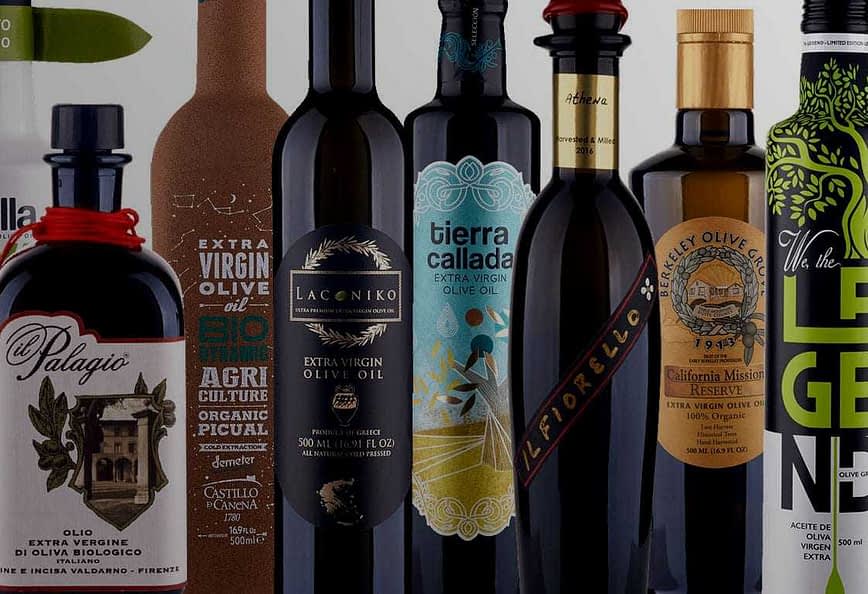 A collection of olive oil bottles with distinct labels and designs arranged in a row. - Olive Oil Times