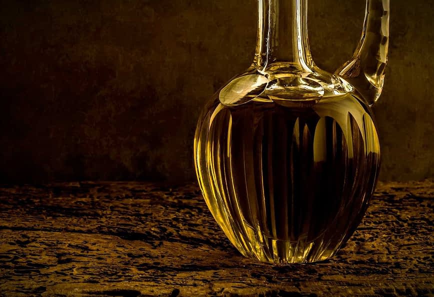 A glass decanter filled with olive oil, featuring a ribbed design and a curved spout. - Olive Oil Times