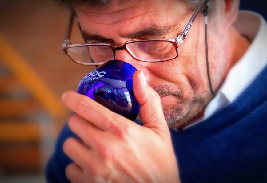 Man with glasses holding a blue glass cup to his nose, appearing to smell it. - Olive Oil Times