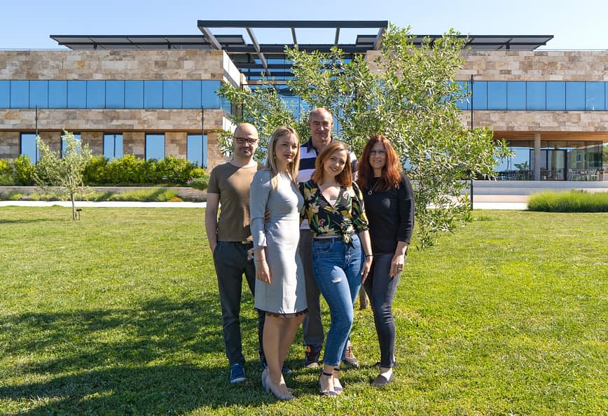 Five individuals standing together outdoors in front of a modern building with greenery. - Olive Oil Times