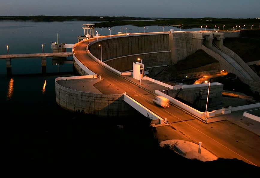 Concrete dam structure with a roadway and lights at dusk, reflecting in the water below. - Olive Oil Times