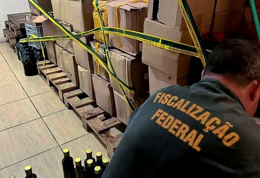 Mit gelbem Klebeband gesicherte Kisten und Flaschen in einem Lagerbereich, wo sie von einer Person inspiziert werden. – Olive Oil Times