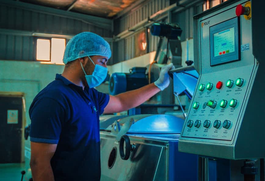 Delavec v modri srajci in mrežici za lase upravlja nadzorno ploščo v industrijskem okolju. - Olive Oil Times