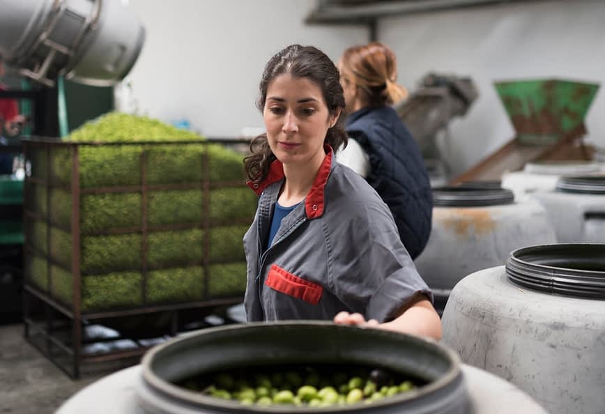 Femme triant des olives dans une usine de production avec de grands conteneurs en arrière-plan. - Olive Oil Times