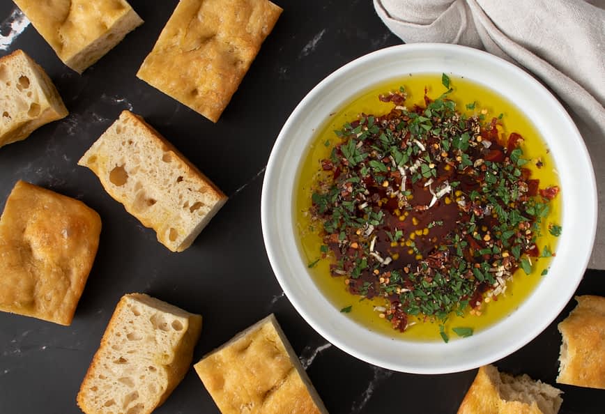 Schüssel mit Olivenöl, gemischt mit Kräutern und Gewürzen, umgeben von Brotstücken auf einer dunklen Oberfläche. - Olive Oil Times