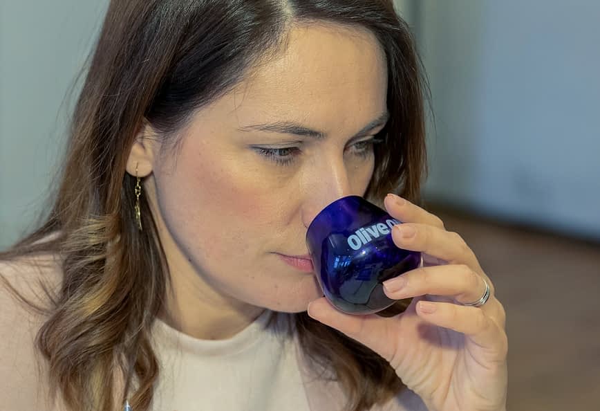Une femme aux cheveux bruns tenant un verre de dégustation bleu étiqueté « Huile d'olive » près de son visage. Olive Oil Times