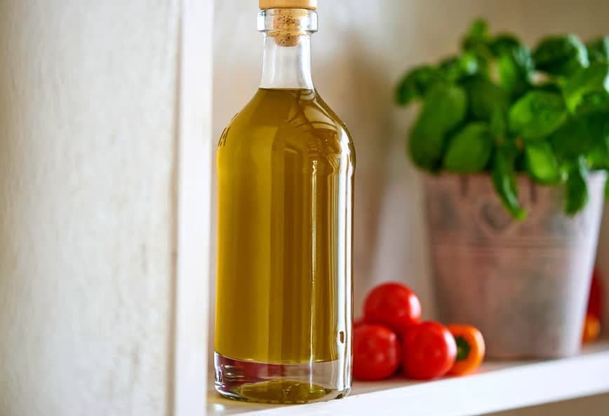 Стеклянная бутылка, наполненная оливковым маслом, стоит на полке рядом с помидорами черри и базиликом. Olive Oil Times