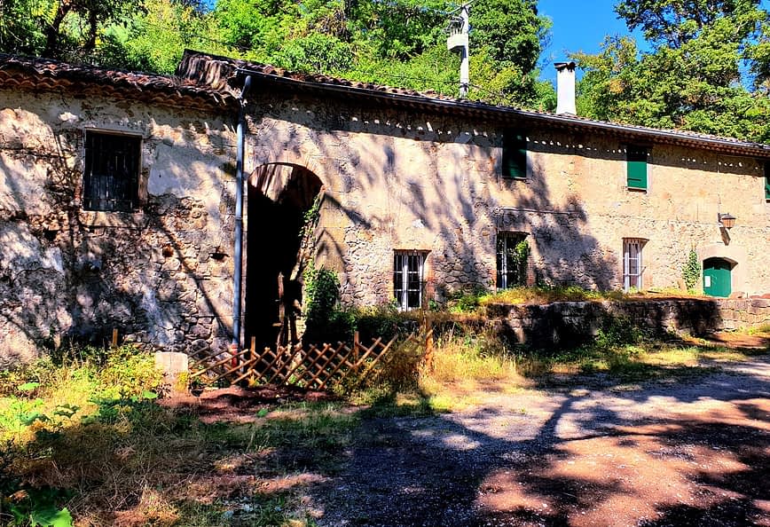 Un edificio rústico de piedra con ventanas y una puerta verdes, rodeado de vegetación. Olive Oil Times