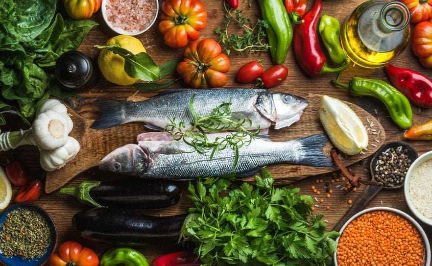 Two whole fish placed on a wooden board surrounded by various vegetables and herbs. - Olive Oil Times