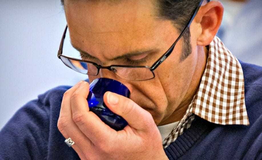 A man wearing glasses and a checkered shirt smelling olive oil from a blue glass cup. - Olive Oil Times