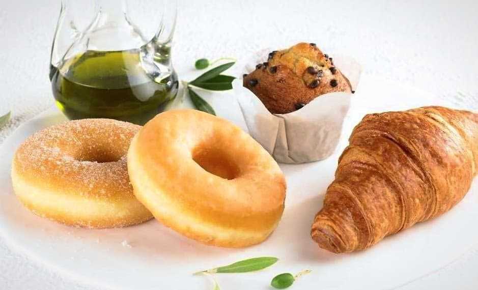 Two sugar-coated donuts, a chocolate chip muffin, and a croissant on a plate with a glass of olive oil. - Olive Oil Times