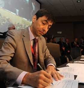 A man in a suit signing documents during a conference with an audience in the background. - Olive Oil Times