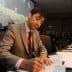 A man in a suit signing documents during a conference with an audience in the background. - Olive Oil Times