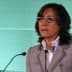 Woman with glasses speaking at a podium during a presentation with a green background. - Olive Oil Times