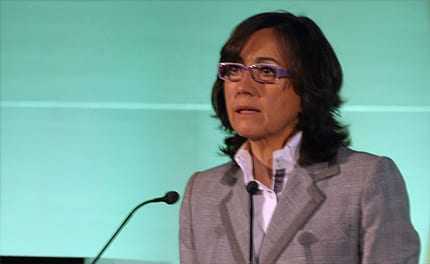 Woman with glasses speaking at a podium during a presentation with a green background. - Olive Oil Times