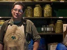 Man wearing an apron standing in an olive shop with jars of olives on shelves behind him. - Olive Oil Times