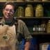 Man wearing an apron standing in an olive shop with jars of olives on shelves behind him. - Olive Oil Times