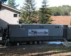 Mobile olive oil processing unit parked next to a building with a vineyard in the background. - Olive Oil Times
