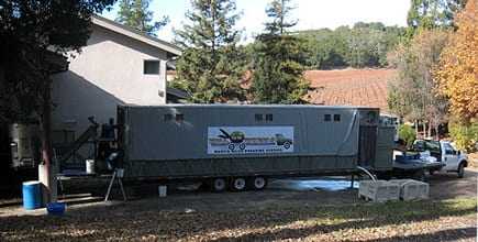 Mobile olive oil processing unit parked next to a building with a vineyard in the background. - Olive Oil Times