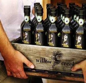 A wooden crate filled with bottles of Willow Creek olive oil held by two people. - Olive Oil Times
