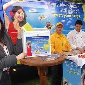 Woman holding a promotional box at an olive oil event with staff and a display table. - Olive Oil Times