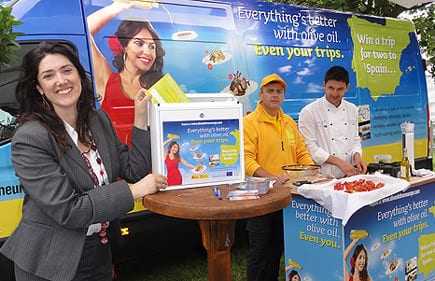 Woman holding a promotional box at an olive oil event with staff and a display table. - Olive Oil Times