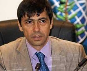 A man wearing a light-colored suit and blue tie sitting at a conference table with a serious expression. - Olive Oil Times