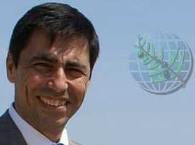 Man wearing a suit and smiling against a clear sky background with a globe graphic. - Olive Oil Times